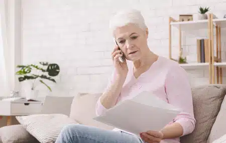 Middle-aged women sitting on her sofa with documents in one hand and speaking on the phone with the other.