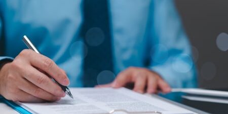 An image of someone wearing a blue shirt with a dark blue tie writing with a pen on a clipboard.