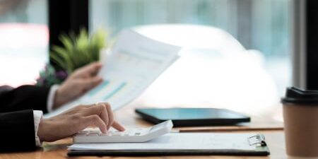 An image of someone typing on their calculator at their desk whilst holding a piece of paper