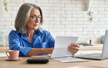 Can-You-Retire-if-You-are-In-Debt-feature An image of a lady who could be at retirement age, sitting at a desk with a coffee, laptop, and reading forms.