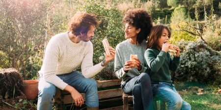 Image of a man and a woman sitting on a bench in the garden with their child, eating sandwiches and smiling