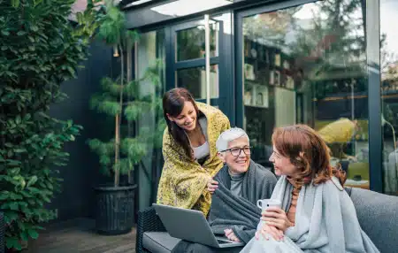 Navigating-Debt-at-Different-Ages-feature Image of a granddaughter, mother, and grandmother all sitting outside, smiling, with a laptop.