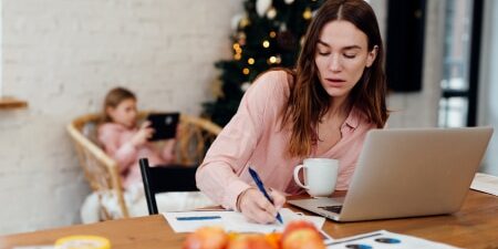 Image of a mother on her laptop writing her budget with her daughter in the background