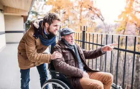 A man using a wheelchair points at something over railings while his son and carer looks on.