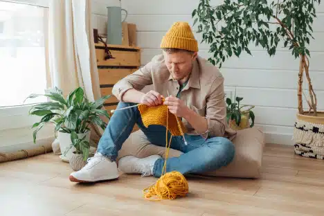 A man in a yellow beanie knitting on the floor in a cozy room.