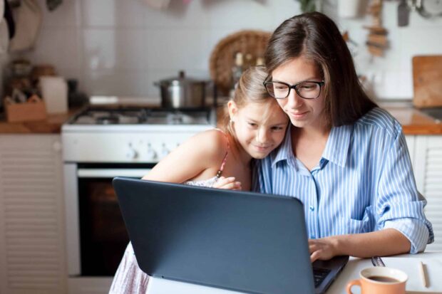 A mother using a laptop while her daughter cuddles up to her.