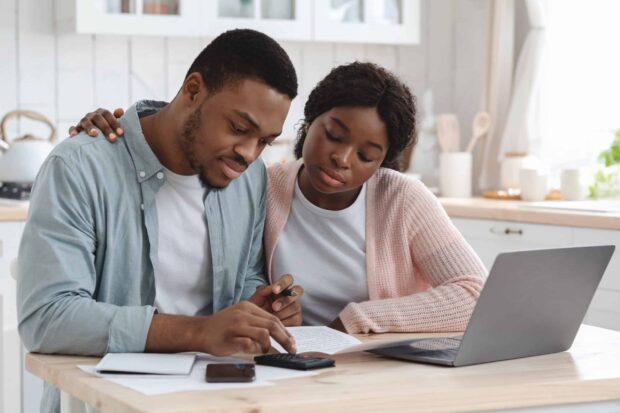 A couple reviews documents together at a kitchen table with a laptop and calculator.