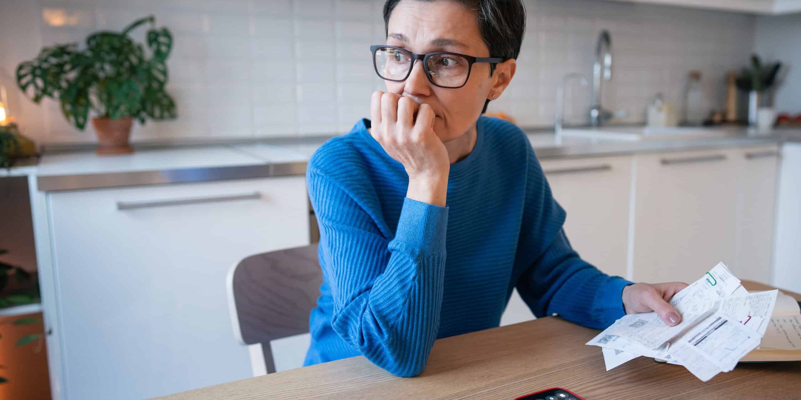 A worried looking woman in a kitchen holding bills, with a calculator app open on her phone.
