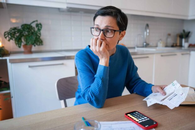 A worried looking woman in a kitchen holding bills, with a calculator app open on her phone.
