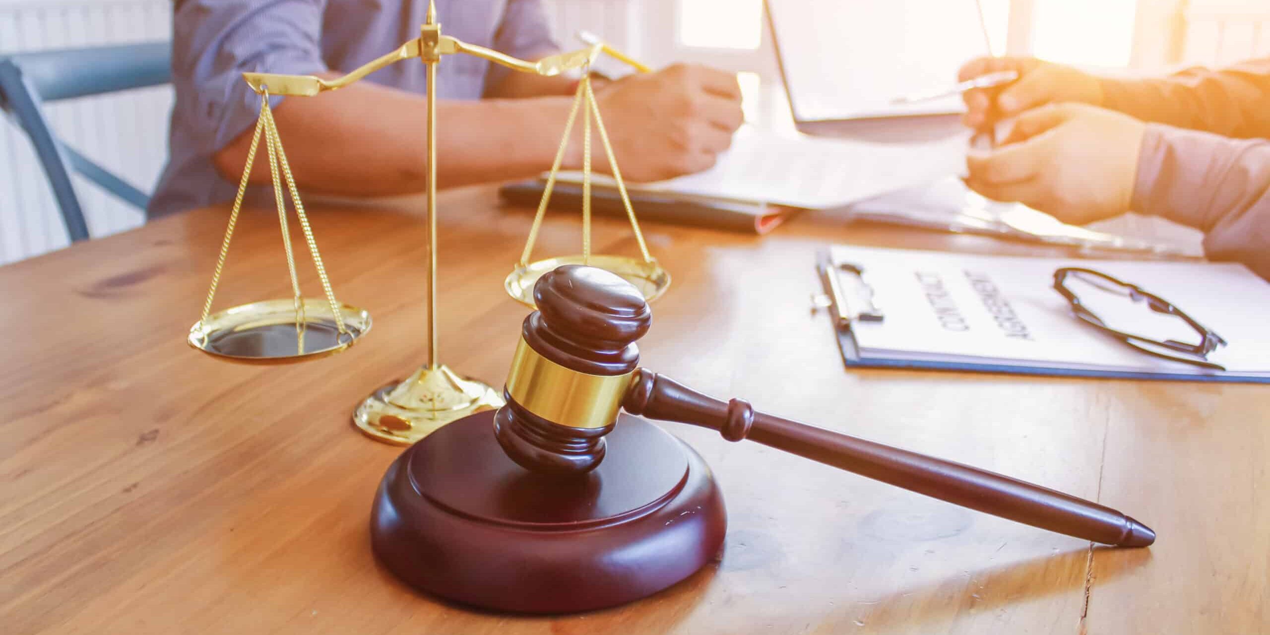 A wooden gavel and golden scales on a desk, with two people reviewing legal documents in the background.