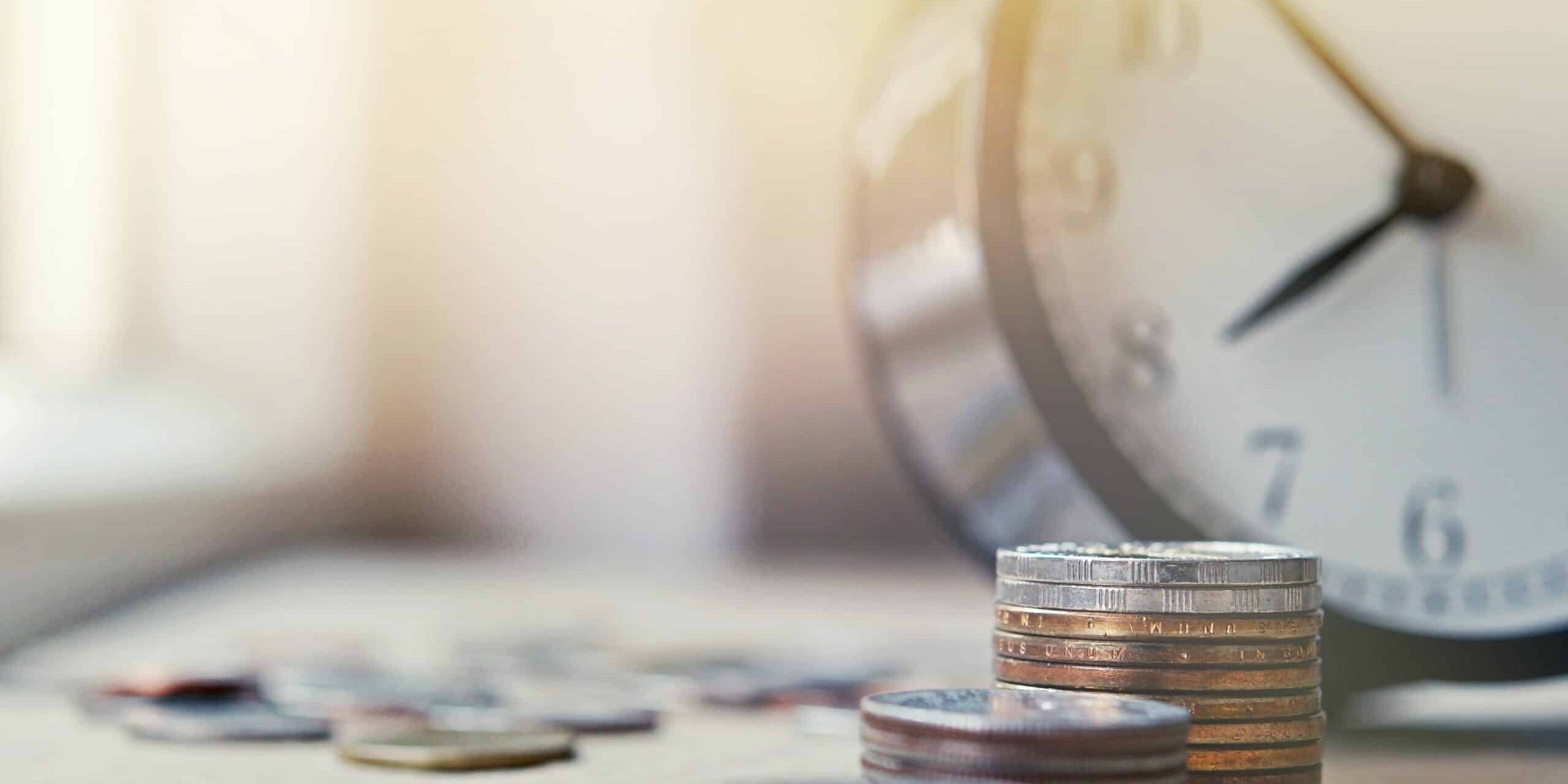 Stacks of coins with a blurred clock in the background, symbolising the concept of time and money.