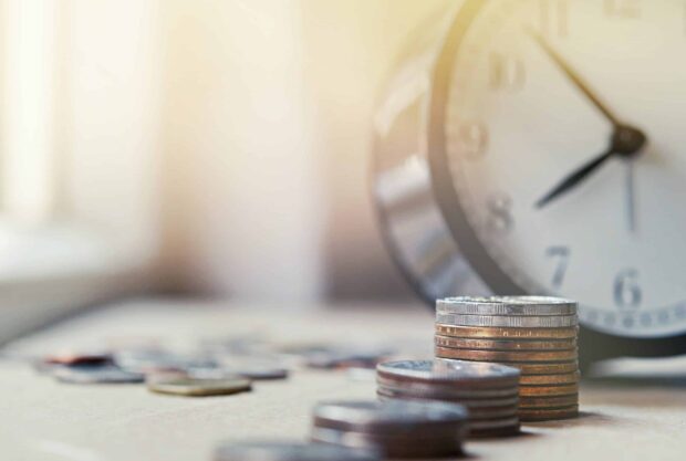 Stacks of coins with a blurred clock in the background, symbolising the concept of time and money.