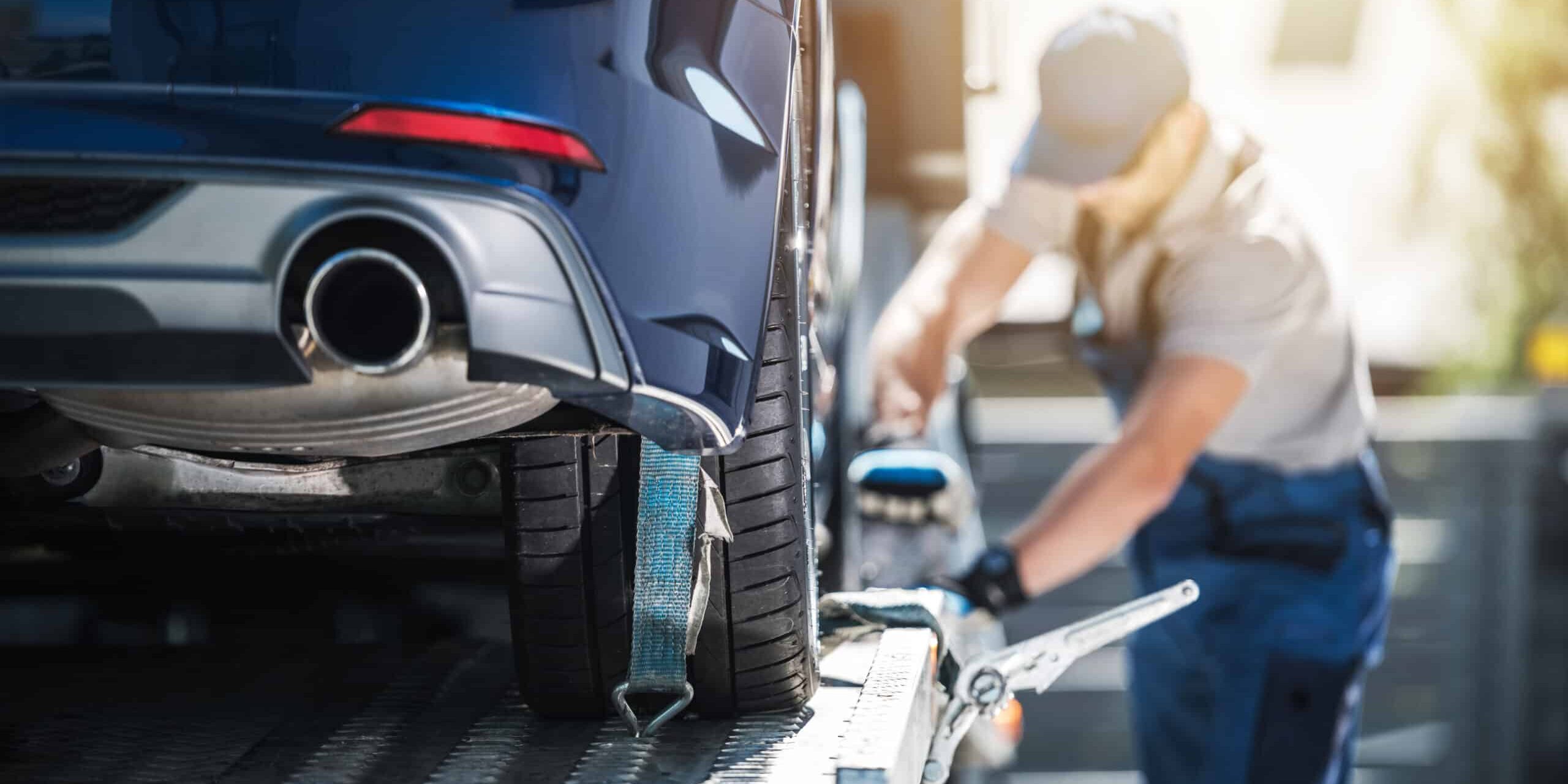 A blue car being secured onto a flatbed tow truck as a worker tightens the strap on the rear tire.