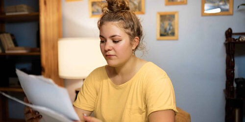 Indoor-image-of-concerned-woman-in-casual-yellow-t-shirt-with-hair-knob-examining-paper-documents-sitting-at-home-in-living-room,-reading-rent-agreement-or-loan-statement-from-bank Indoor image of concerned woman in casual yellow t-shirt with hair knob examining paper documents sitting at home in living-room, reading rent agreement or loan statement from bank