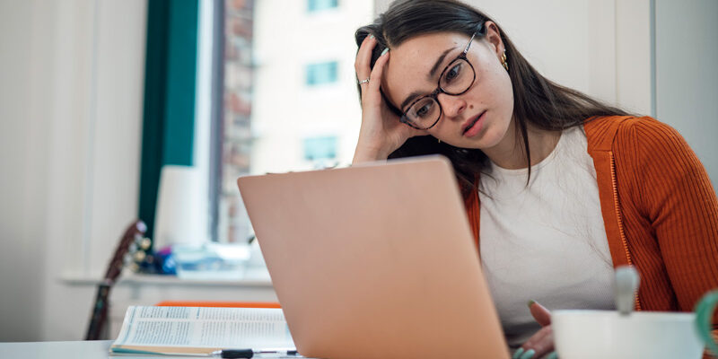 Front view of a young woman wearing casual clothing sitting at a desk using a laptop to study, she is a university student. She is feeling stressed and she has her head in her hand.