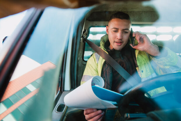Mid-adult manual worker is sitting in his van, looking through paperwork while speaking to someone on the phone.